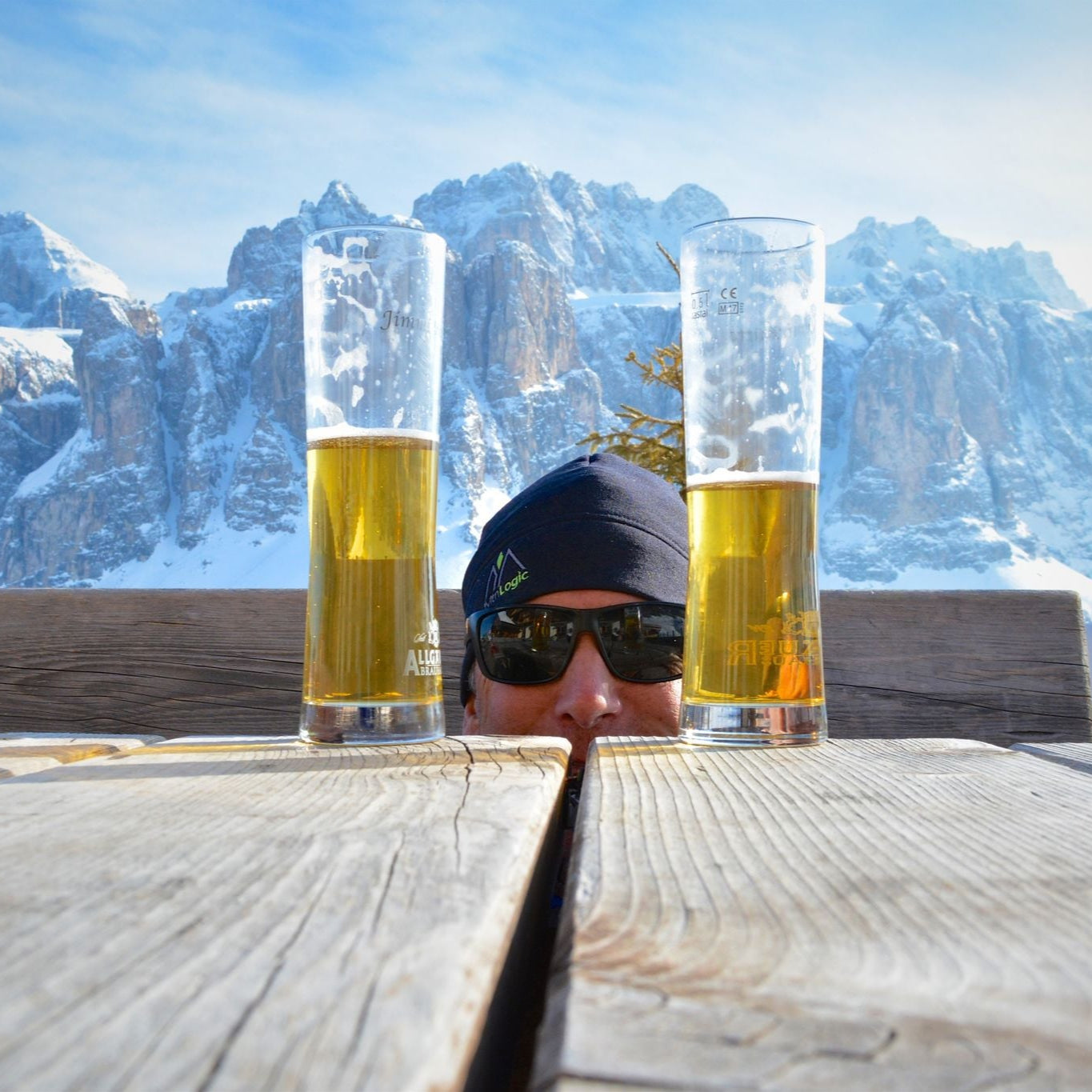 Person wearing sunglasses with two beer glasses in front of a snowy mountain landscape